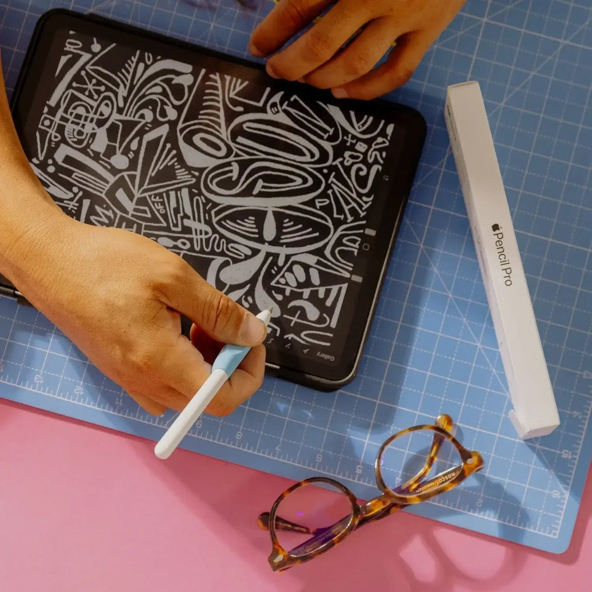 Ręce rysujące na tabletu z długopisem, obok okulary i pudełko Pencil Pro na niebieskiej matce